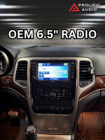 Interior view of a 2011-2013 Jeep Grand Cherokee featuring the factory-installed OEM 6.5-inch radio. The screen displays the Jeep logo with standard radio controls and navigation options below. This image serves as a "before" comparison prior to upgrading to the Sony XAV-9500ES radio with Prolific Audio's DIY kit.