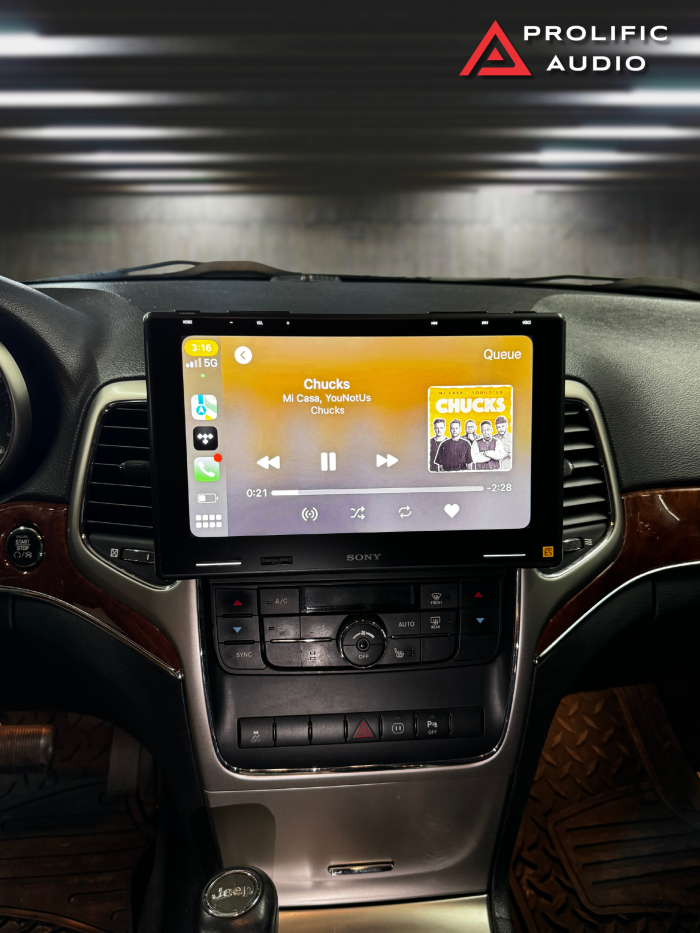 Interior view of a 2011-2013 Jeep Grand Cherokee featuring the Sony XAV-9500ES radio installed with Prolific Audio's DIY kit, running Apple CarPlay. The screen displays a music player interface with the song "Chucks" by Mi Casa and YouNotUs, showing album art and playback controls. The installation maintains the factory HVAC controls below for a seamless and integrated user experience.