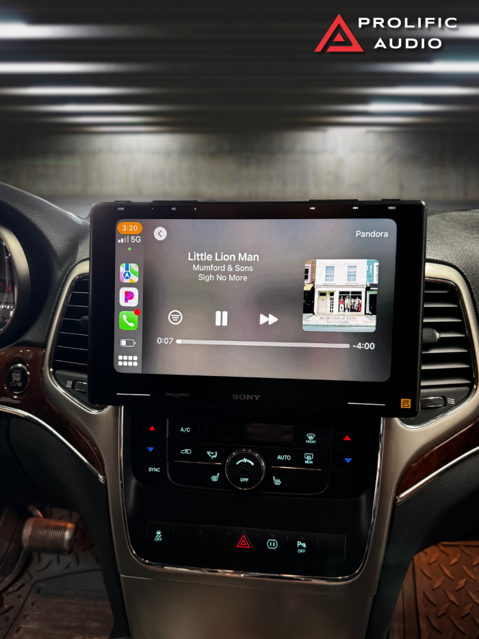 Interior view of a 2011-2013 Jeep Grand Cherokee featuring the Sony XAV-9500ES radio installed using Prolific Audio's DIY kit. The screen displays a music player interface on Apple CarPlay, showing "Little Lion Man" by Mumford & Sons from the album "Sigh No More," along with album art and playback controls. The custom bezel ensures a seamless fit while retaining the factory HVAC controls below for a professional and integrated look.