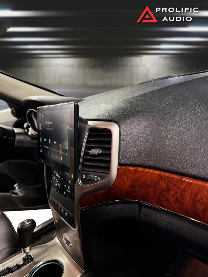 Side view of the interior of a 2011-2013 Jeep Grand Cherokee showcasing the Sony XAV-9500ES radio installed using Prolific Audio's DIY kit. The image highlights the clean integration of the radio with the dashboard, utilizing a custom bezel that seamlessly blends with the woodgrain trim, while maintaining the factory HVAC controls and displaying a music interface on the screen.