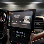 Side-angle view of the interior of a 2011-2013 Jeep Grand Cherokee showcasing the Sony XAV-9500ES radio installed using Prolific Audio's DIY kit. The screen is running Apple CarPlay, displaying the Pandora music interface with the song "Little Lion Man" by Mumford & Sons from the album "Sigh No More," including album art and playback controls. The custom bezel ensures a clean and professional integration with the dashboard, retaining the factory HVAC controls below.
