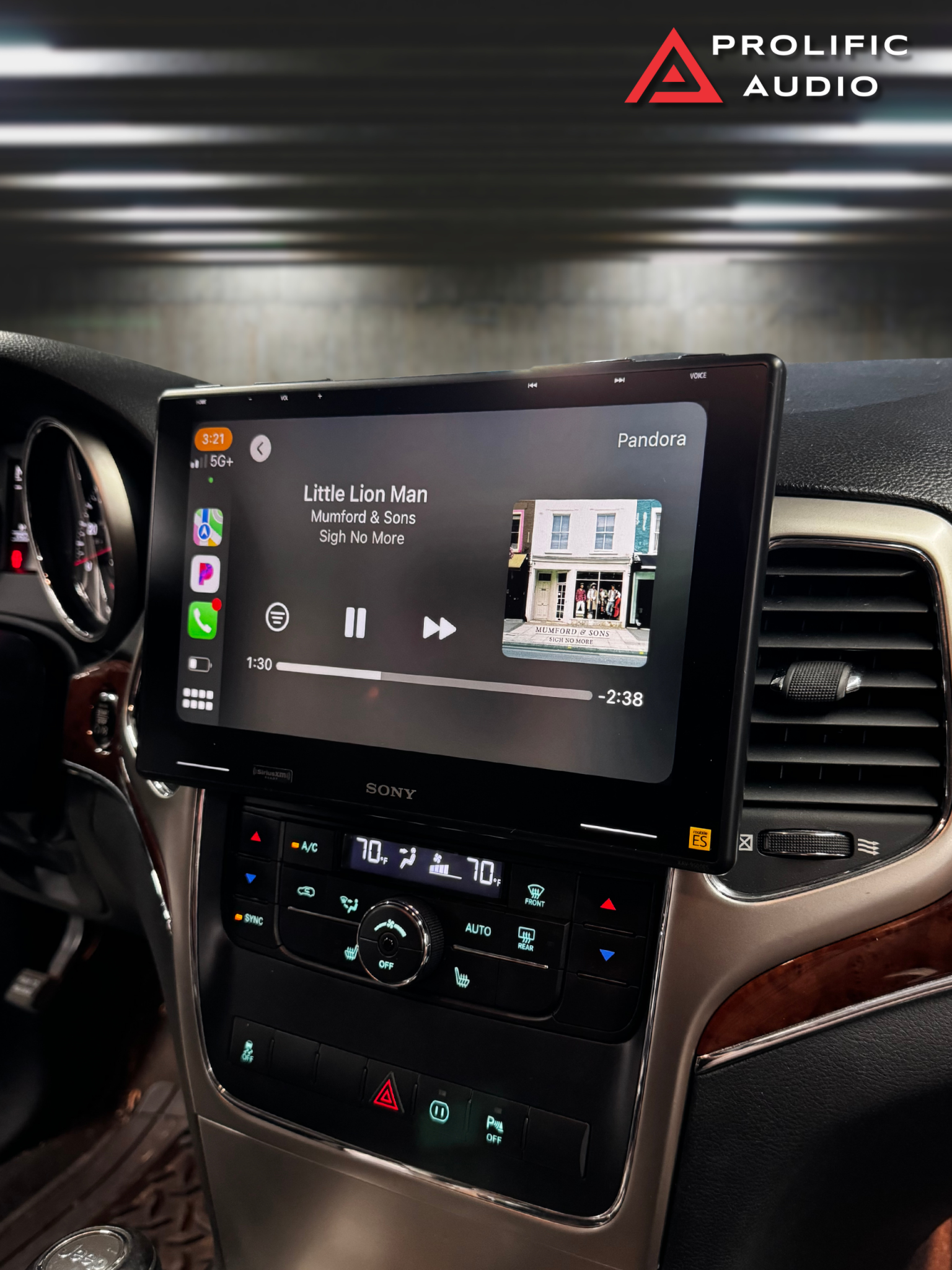 Sony XAV-9500ES Music Playback Interface Side-angle view of the interior of a 2011-2013 Jeep Grand Cherokee showcasing the Sony XAV-9500ES radio installed using Prolific Audio's DIY kit. The screen is running Apple CarPlay, displaying the Pandora music interface with the song "Little Lion Man" by Mumford & Sons from the album "Sigh No More," including album art and playback controls. The custom bezel ensures a clean and professional integration with the dashboard, retaining the factory HVAC controls below.
