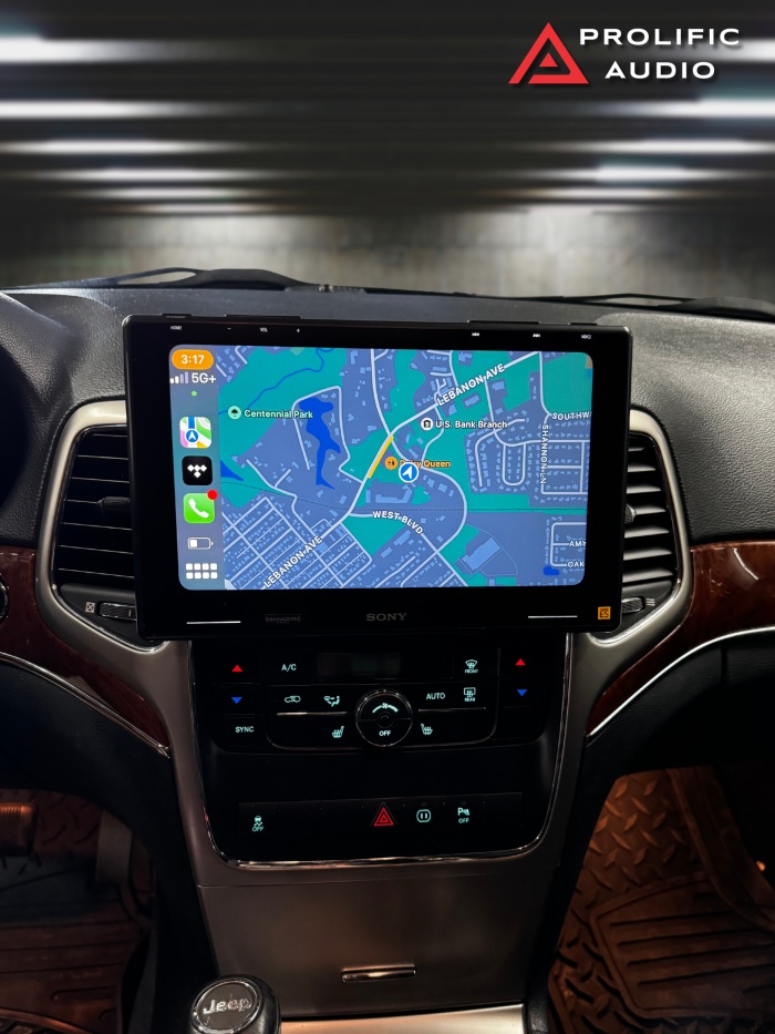 Interior view of a 2011-2013 Jeep Grand Cherokee with the Sony XAV-9500ES radio installed using Prolific Audio's DIY kit, running Apple CarPlay. The screen displays a navigation map showing local streets and landmarks, providing turn-by-turn directions. The setup integrates seamlessly with the factory HVAC controls below for a clean and functional appearance.