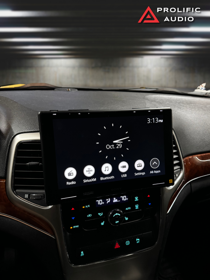 Interior view of a 2011-2013 Jeep Grand Cherokee with the Sony XAV-9500ES radio installed using Prolific Audio's DIY kit. The radio displays the home screen featuring a clock with the date October 29, alongside menu icons for radio, SiriusXM, Bluetooth, USB, settings, and all apps. The installation maintains the factory HVAC controls below for a seamless and integrated look.