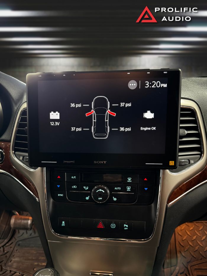 Interior view of a 2011-2013 Jeep Grand Cherokee featuring the Sony XAV-9500ES radio installed using Prolific Audio's DIY kit. The screen displays vehicle diagnostics, including tire pressures for each wheel, battery voltage, and engine status. The custom bezel ensures a seamless fit, integrating the radio cleanly into the dashboard while retaining the factory HVAC controls below.