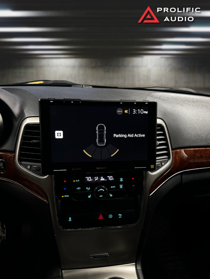 Interior view of a 2011-2013 Jeep Grand Cherokee featuring the Sony XAV-9500ES radio installed with Prolific Audio's DIY kit. The screen displays the vehicle's parking aid system with visual proximity alerts, seamlessly integrated into the dashboard while retaining full functionality of the factory HVAC controls below.