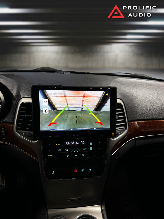 Interior view of a 2011-2013 Jeep Grand Cherokee with the Sony XAV-9500ES radio installed using Prolific Audio's DIY kit. The screen displays a rearview camera feed with parking guide lines, seamlessly integrated into the dashboard for a professional and functional appearance, while retaining the factory HVAC controls below.